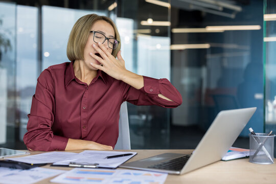 Businesswoman feeling tired and stressed, yawning during a long workday, experiencing burnout and fatigue at her office desk with a laptop and documents
