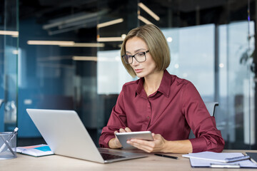 Confident businesswoman with eyeglasses working diligently on a laptop and holding a digital tablet at a light wooden desk, focusing on tasks in a contemporary office environment