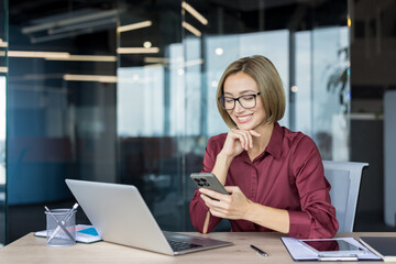 Happy businesswoman wearing eyeglasses smiles, looking at her smartphone while sitting at a desk with a laptop in a contemporary office environment, engaging with digital content