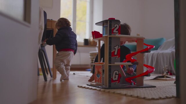 Toddler exploring toy car garage while older sibling plays nearby, independent discovery beside shared play, early childhood curiosity autonomy and gentle family presence at home