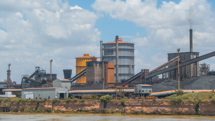 Obraz premium Industrial landscape of a large factory complex with silos, conveyor belts, and smokestacks lining the bank of a river