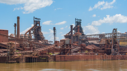 Industrial landscape of a massive, rust-colored mining complex by a muddy river under a blue sky with white clouds