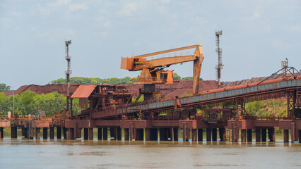 Industrial port facility featuring a large orange bulk materials crane, conveyor belt, and massive ore piles against a calm sky
