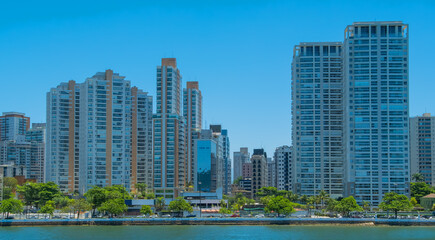 Modern coastal skyline with high-rise residential and commercial buildings towering over a lush green waterfront, reflecting a bright, sunny day