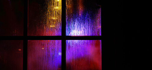 Fototapeta premium A dark window pane with rain droplets at night. Blurred yellow and blue festive christmas lights through the wet glass covered with condensate drops. Abstract background.