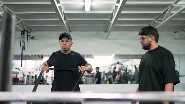 Young man with tattoos performing an upright row with a resistance band while his coach supervises