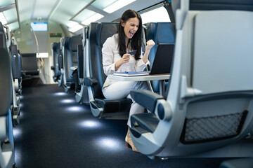 This image captures a joyful woman celebrating a success while using her laptop and bank card on a train, showcasing the happiness derived from digital travel pay