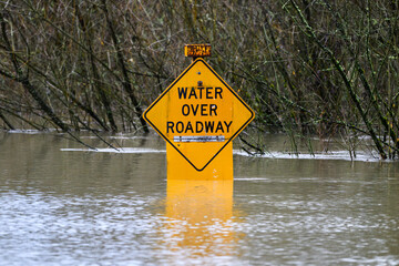 Flooded muddy river water surrounds yellow road sign with message water over roadway