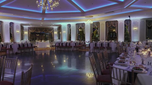 Empty Hotel Ballroom With Blue Uplighting, Round Tables Dressed In White Linen, Chiavari Chairs, Elevated Head Table With Backdrop, Polished