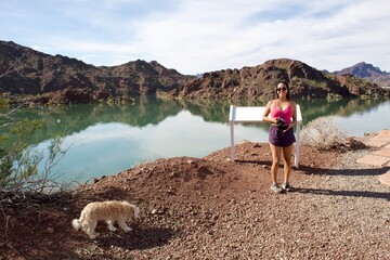 Fototapeta premium Asian girl and small fluffy white dog hiking at Bill Williams River with beautiful mountains reflecting on calm water in Arizona