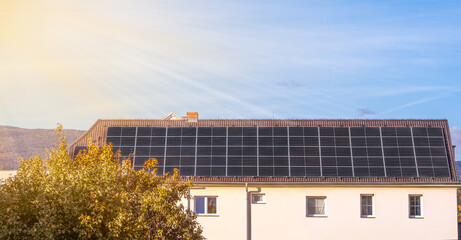 Obraz premium Modern residential building with solar panels installed on the roof, capturing clean renewable energy from the sun. Eco friendly home with photovoltaic system, blue sky, warm sunlight, and trees in