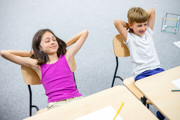 School children relaxing at desks after lesson, leaning back with hands behind head. Education, school routine, learning break, classroom lifestyle and childhood concept