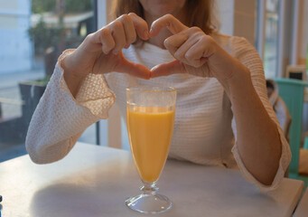 A woman forms a heart shape with her hands over a glass of fresh orange juice while sitting at a coffe table