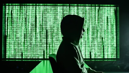 A hooded silhouette at a computer, bathed in cascading green code evoking cyberpunk hacking moodscape - Powered by Adobe