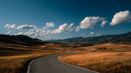 Winding asphalt road disappears into golden rolling hills beneath a bright blue sky with scattered white clouds