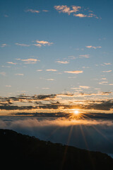A breathtaking sunrise illuminates the horizon above a sea of clouds, casting a warm glow against a backdrop of blue sky and scattered clouds.