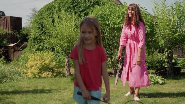 Caucasian Mother And Daughter Greeting In Sunlit Garden, Mother In Pink Dress Welcomes Young Girl Holding Racket, Lush Hedges And Lawn, Tender Hug And Smiling Faces, Joyful Family Moment