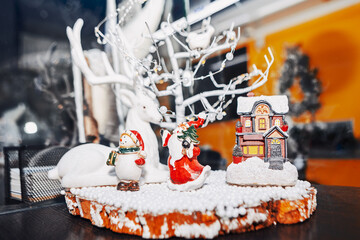 Festive Christmas scene features white reindeer, snowman, Santa, and fairy house nestled in snowy decor. Warm lights and a blurred wreath enhance the cozy, celebratory holiday atmosphere