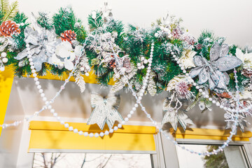 Festive garland of green pine branches hangs above window, adorned with silver poinsettias, pinecones, berries and sparkling beads. Elegant arrangement glows with seasonal charm and decorative detail