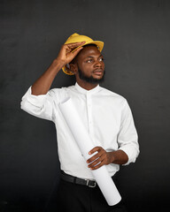 A man wearing a white shirt and yellow safety helmet holds a rolled construction plan while...