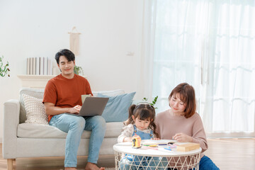 Asian mother & daughter concentrating on coloring homework father works remotely on laptop in cozy living room. Heartfelt scene illustrating modern family work-life balance and remote work.