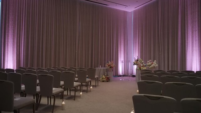 Aisle Lined Chairs With Purple Uplighting And Candles Leading To Floral Altar Empty Boutique Ballroom Framed By Heavy Drapery, Soft