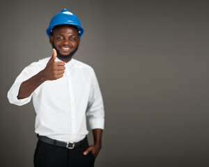 A young African engineer wearing a blue safety helmet and white shirt stands in a studio giving a thumbs up gesture, smiling confidently while expressing approval in a professional construction 