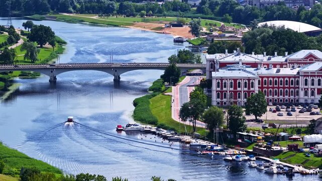 Aerial view of Jelgava Palace beside the Lielupe River, with boats moving across the water and a historic landmark surrounded by green parks and bridges. A scenic Latvian city landscape.