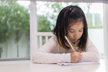 Focused Asian girl sitting at a white table, intently writing in a notebook with a pencil while...