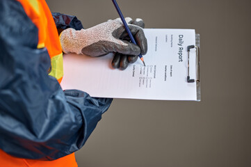 Close up of an African construction worker wearing gloves and a reflective safety vest, holding a clipboard and writing on a printed daily report form against a plain studio background.