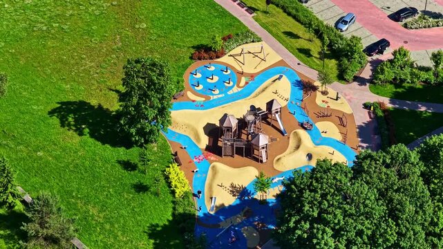 Aerial view of a colorful modern playground with sand zones, blue pathways, climbing structures, and slides, surrounded by green trees and nearby parking areas on a sunny day.