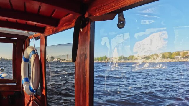 Sunlit river waves reflect through a boat&rsquo;s clear side panel while warm wooden beams and a life ring frame the shoreline in the background, creating a calm nautical travel moment.