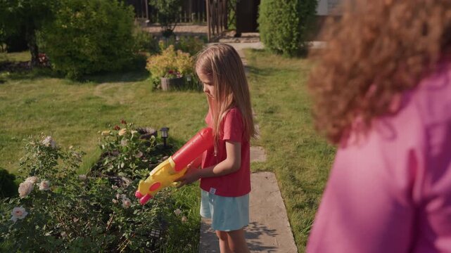 Youthful Flower Play, Girl Enjoys Spraying Garden Blooms Under Bright Sunny Sky While Adult Observes, Young Girl Mischievously Sprays Flowers In Sunny Garden As Adult Watches Nearby From Patio