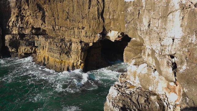 Slow motion video of turquoise large and foamy waves hitting a large and steep cliff bank on Portugal coastline called Boca do Inferno or Devils mouth while sun shines during day time. Tourism spot.