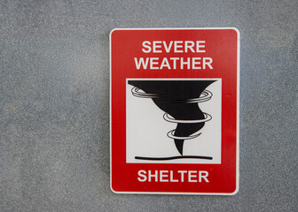 A red sign for a Severe Weather Shelter against a gray wall with a graphic of a tornado or hurricane