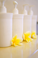 Soft focus image featuring white pump bottles arranged in a row with fresh yellow plumeria flowers at the base. Clean, spa-like scene ideal for beauty, skincare, and wellness branding.