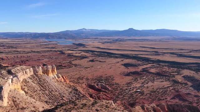 Drone footage glides above red rock cliffs into wide desert plains, revealing layered badlands, sparse vegetation, a distant reservoir, and Cerro Pedernal rising prominently along the horizon.