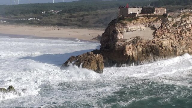 Beautiful aerial drone shot of big waves breaking on rocks and cliffs on a day with giant waves in Nazar&eacute;, Portugal, Europe. Farol da Nazare lighthouse and wind turbines visible