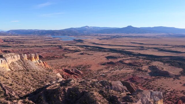 Drone footage above red rock cliffs into broad desert plains, revealing layered badlands, sparse vegetation, a distant lake, and Cerro Pedernal rising along the horizon beneath clear blue skies.