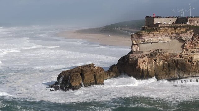 Aerial drone shot of big waves breaking on rocks and cliffs on a day with giant waves in Nazar&eacute;, Portugal, Europe. Farol da Nazar&eacute; lighthouse and wind turbines