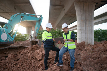 Two construction engineers reviewing digital plans on a laptop at infrastructure site, standing on...