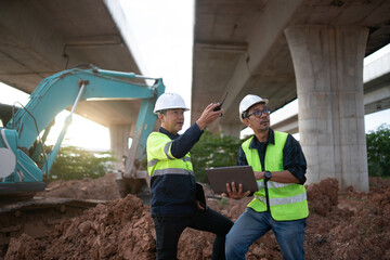 Two engineers standing on soil mound discussing infrastructure plans beside excavator, highlighting...