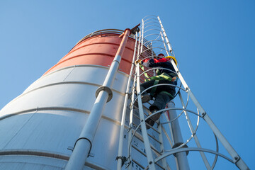 Maintenance worker climbing an enclosed ladder attached to a metal silo, secured with safety gear, representing regulated access and high-altitude industrial work.
