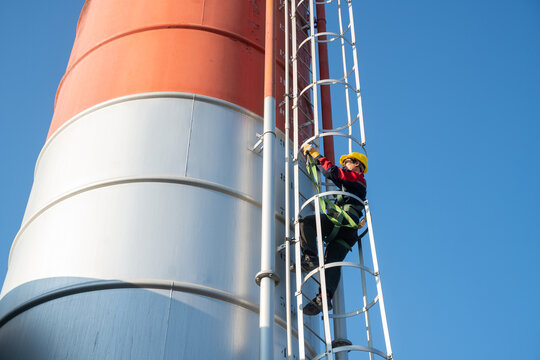 Low-angle view of a worker ascending a safety cage ladder on a cylindrical industrial tank, highlighting scale, engineering structure, and fall protection systems.