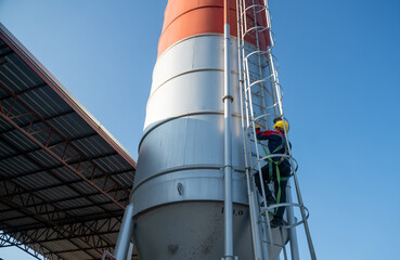 Worker climbing a vertical ladder on an outdoor industrial silo under clear blue sky, emphasizing maintenance responsibility and occupational safety.