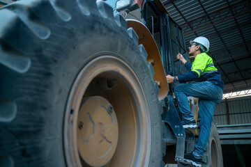 Professional industrial worker wearing safety helmet and high-visibility uniform climbing heavy construction machinery inside a factory warehouse