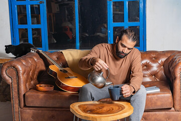 Person pouring tea from metal teapot beside guitar and cat in cozy room