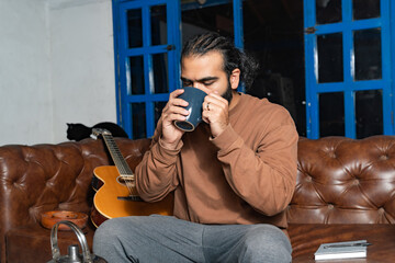 Person drinking tea beside guitar and cat on leather couch in relaxed setting