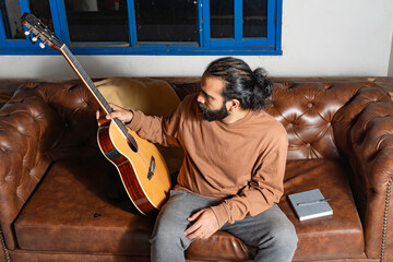 Person examining acoustic guitar neck while seated on leather couch with notebook