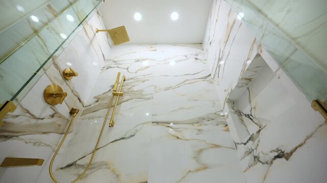 POV of opening the glass doors of the shower in the luxury bathroom with elegant gold fixtures. The marble-style wall tile adds a high-end, sophisticated look.